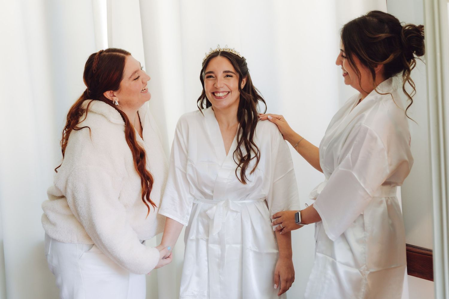 Bridesmaids in white robes helping bride with hair during wedding morning prep.