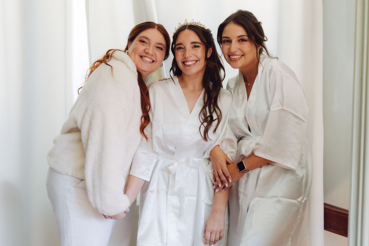 Bride and two bridesmaids in white robes smiling together before wedding ceremony.