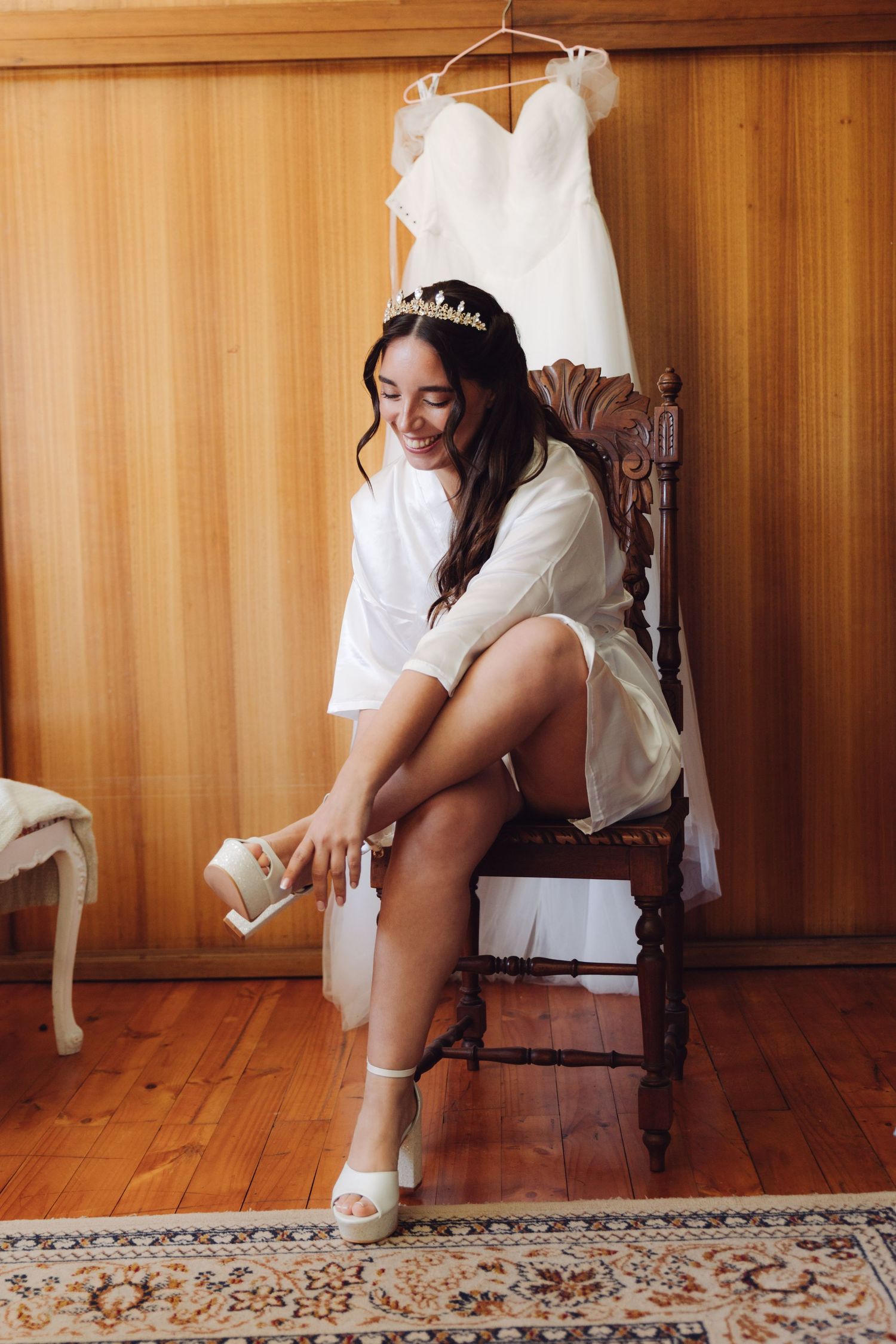 Bride in white robe wearing tiara putting on wedding shoes before ceremony.