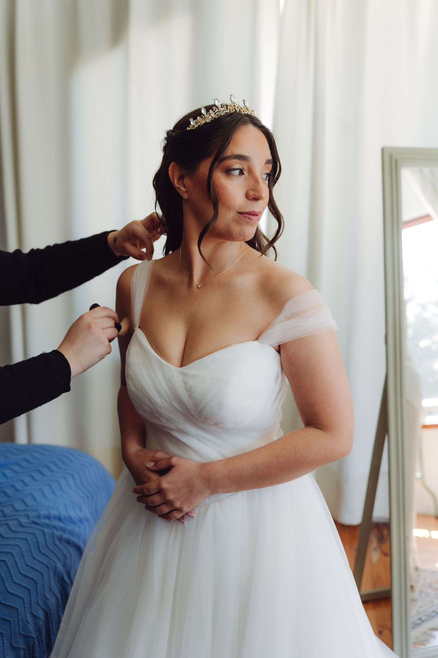 Bride in white off-shoulder wedding gown with tiara being dressed by assistant.