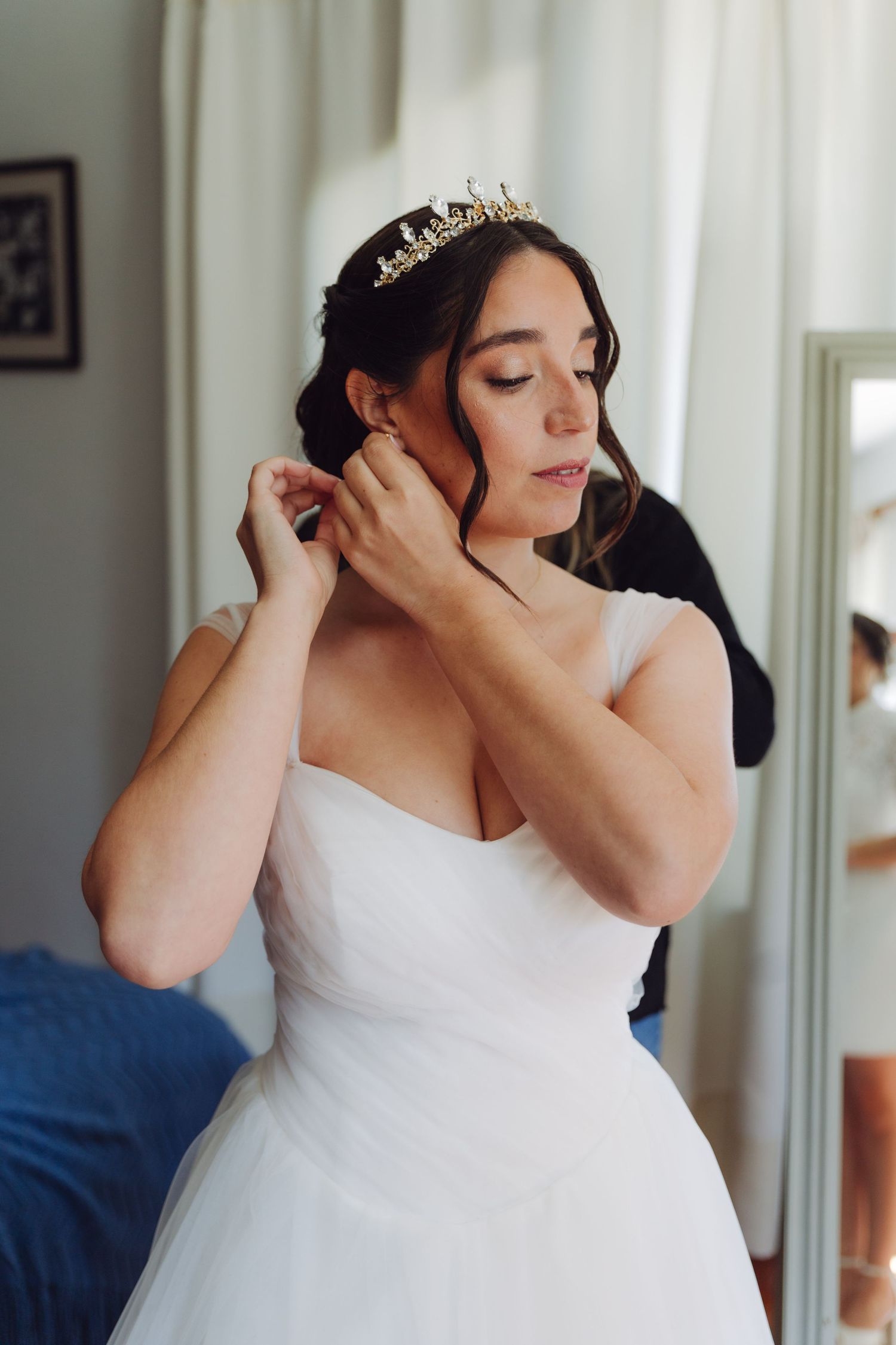 Bride in white gown and gold tiara putting on earrings while getting ready for wedding.