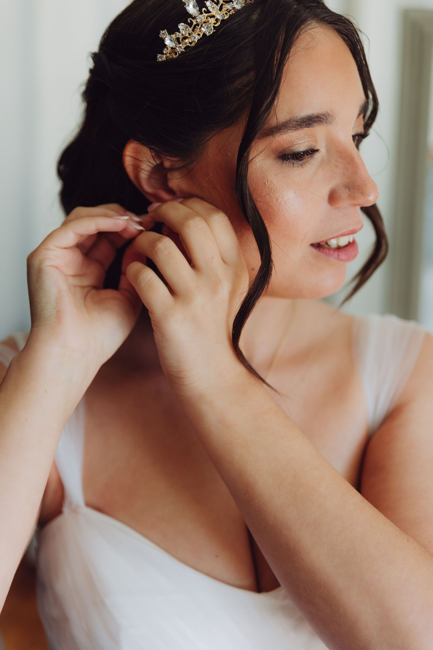 Beautiful bride wearing gold tiara putting on earrings in wedding dress.