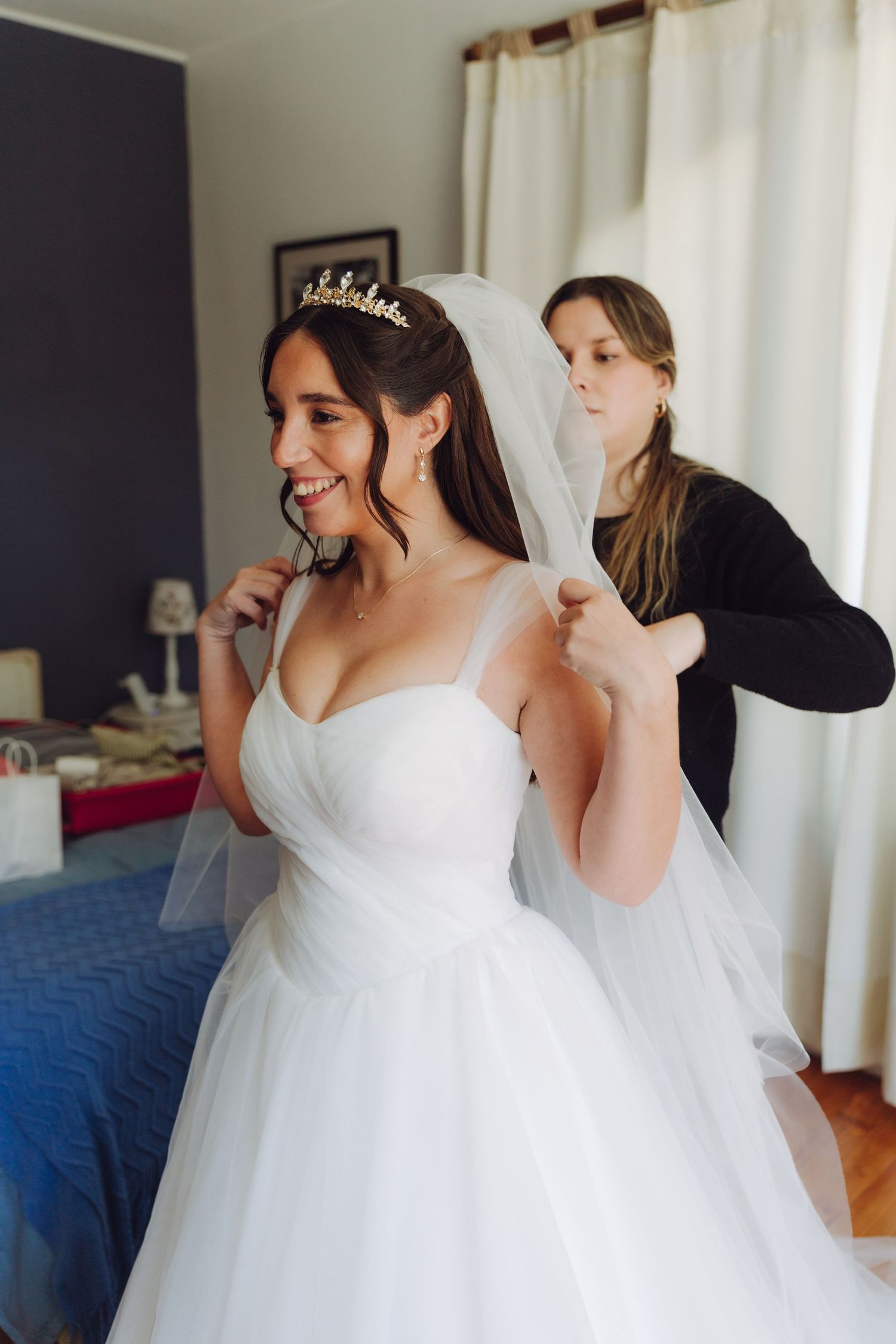 Smiling bride in white wedding gown with tiara having veil attached by assistant.