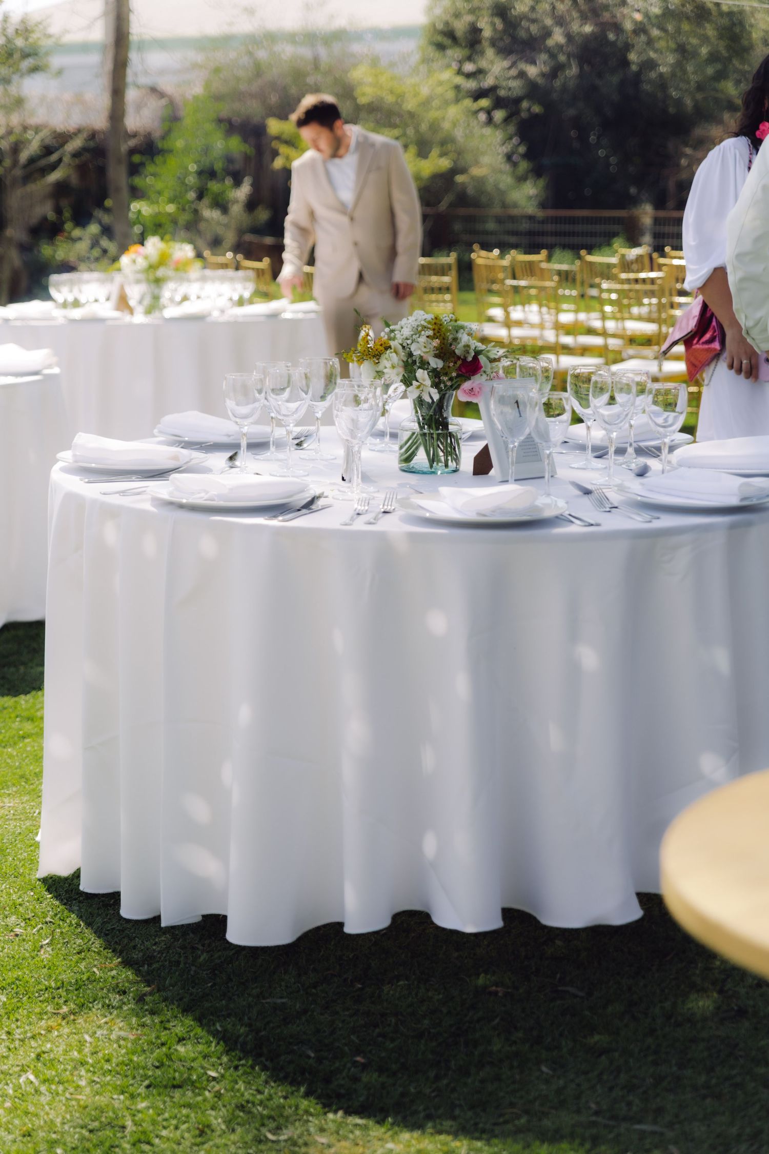 Elegant outdoor wedding reception tables with white linens, glassware, and floral centerpieces on green lawn.