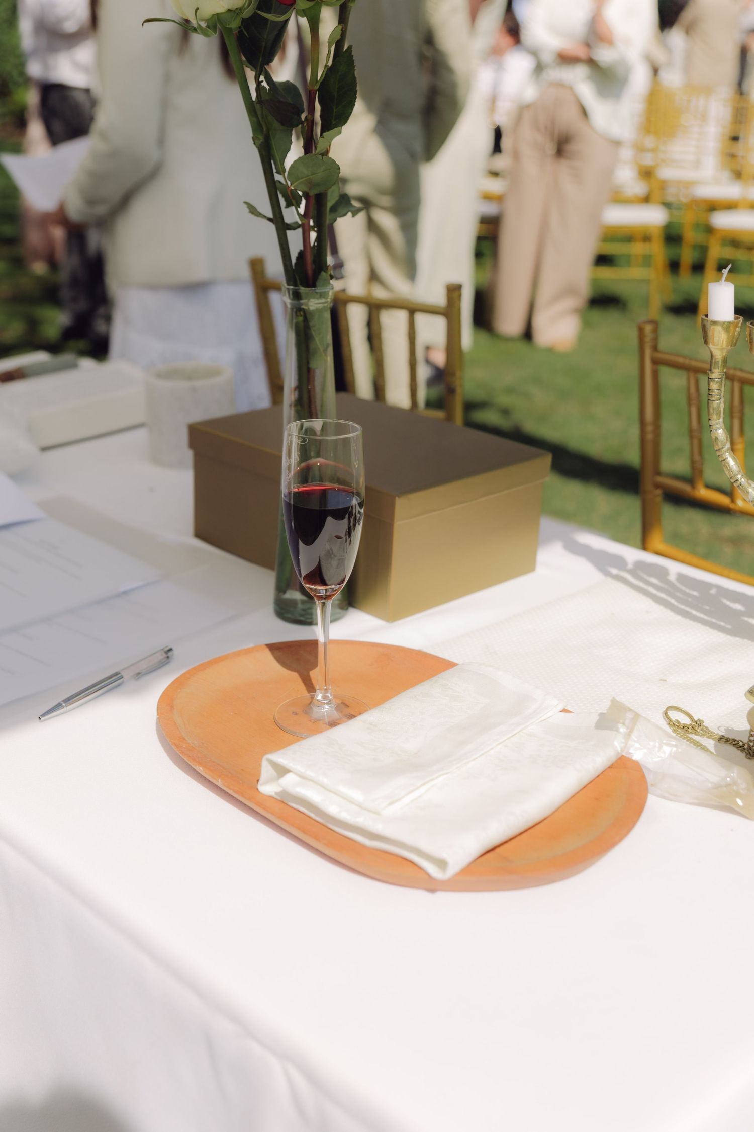Wedding ceremony table with champagne flute, white napkin on terracotta plate, rose vase, and gold gift box.