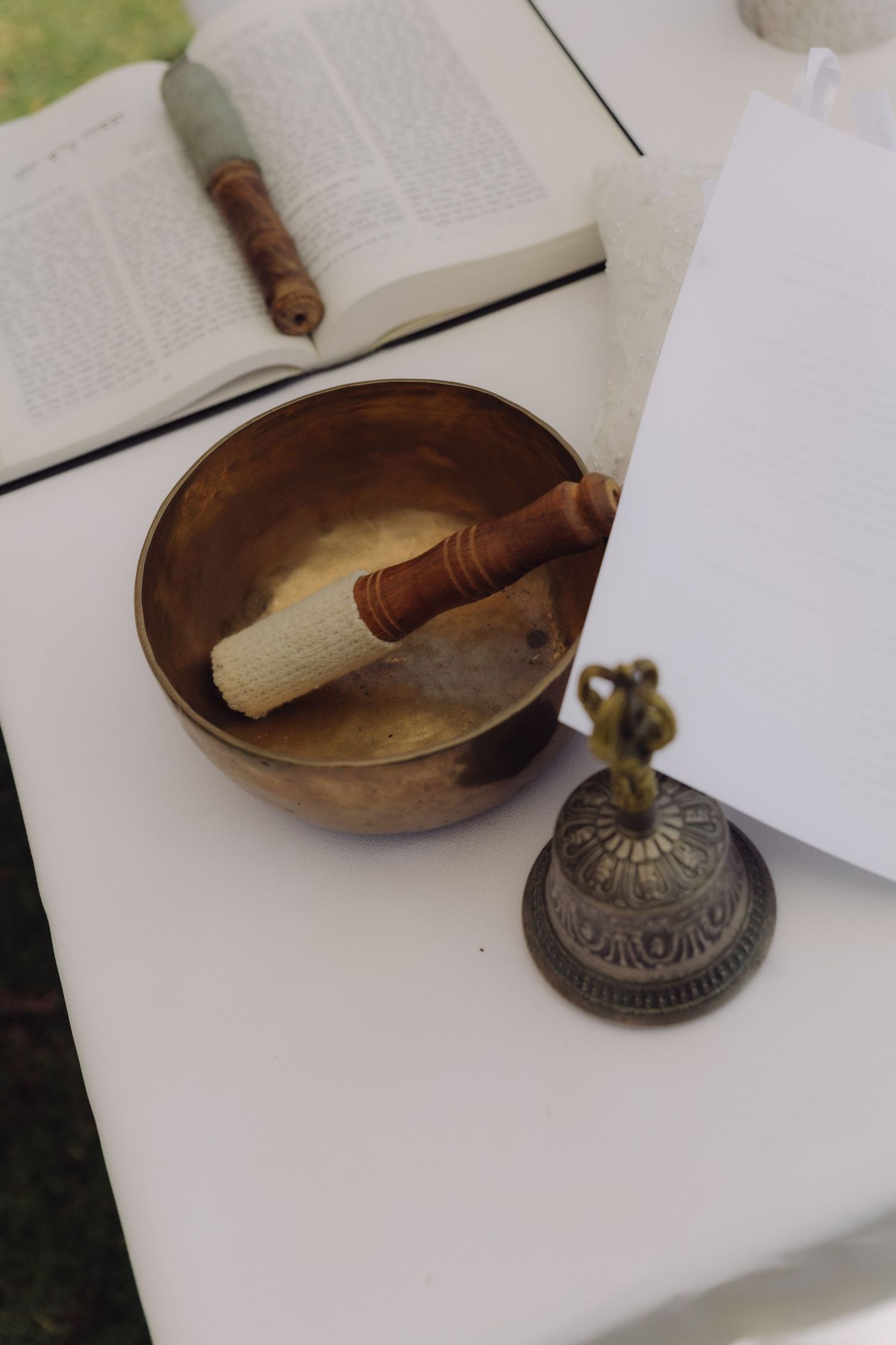 Tibetan singing bowl with mallet, decorative bell, and open book on white table for outdoor wedding ceremony.