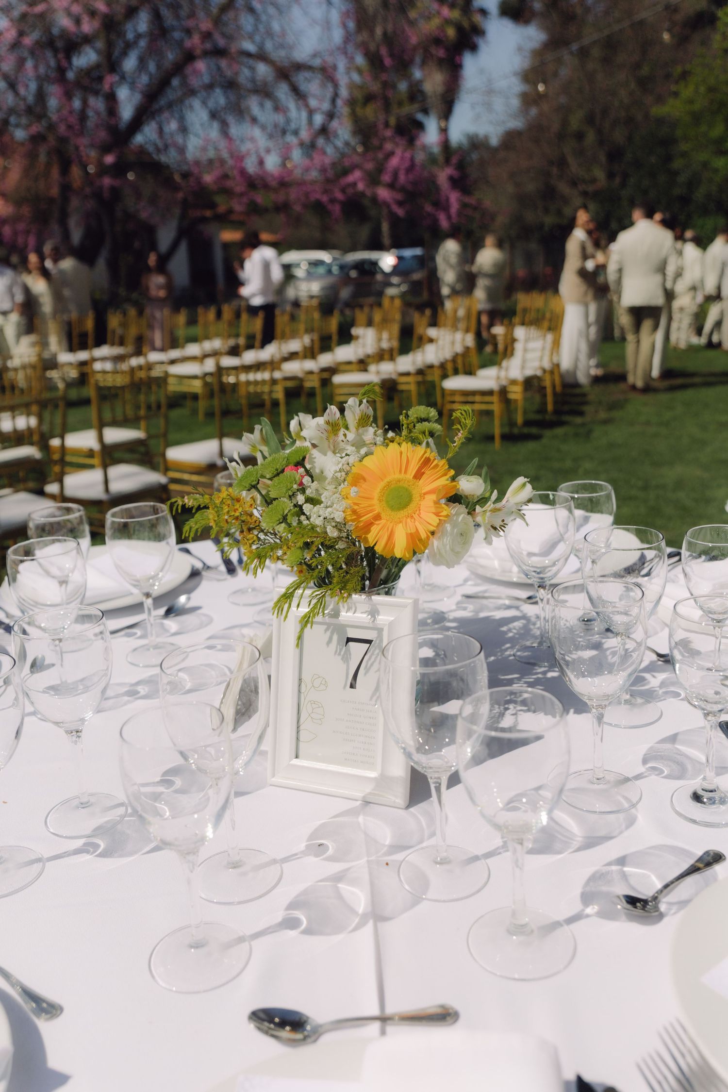 Outdoor wedding reception table with white linen, yellow gerbera daisy centerpiece, glassware, and table number 7.