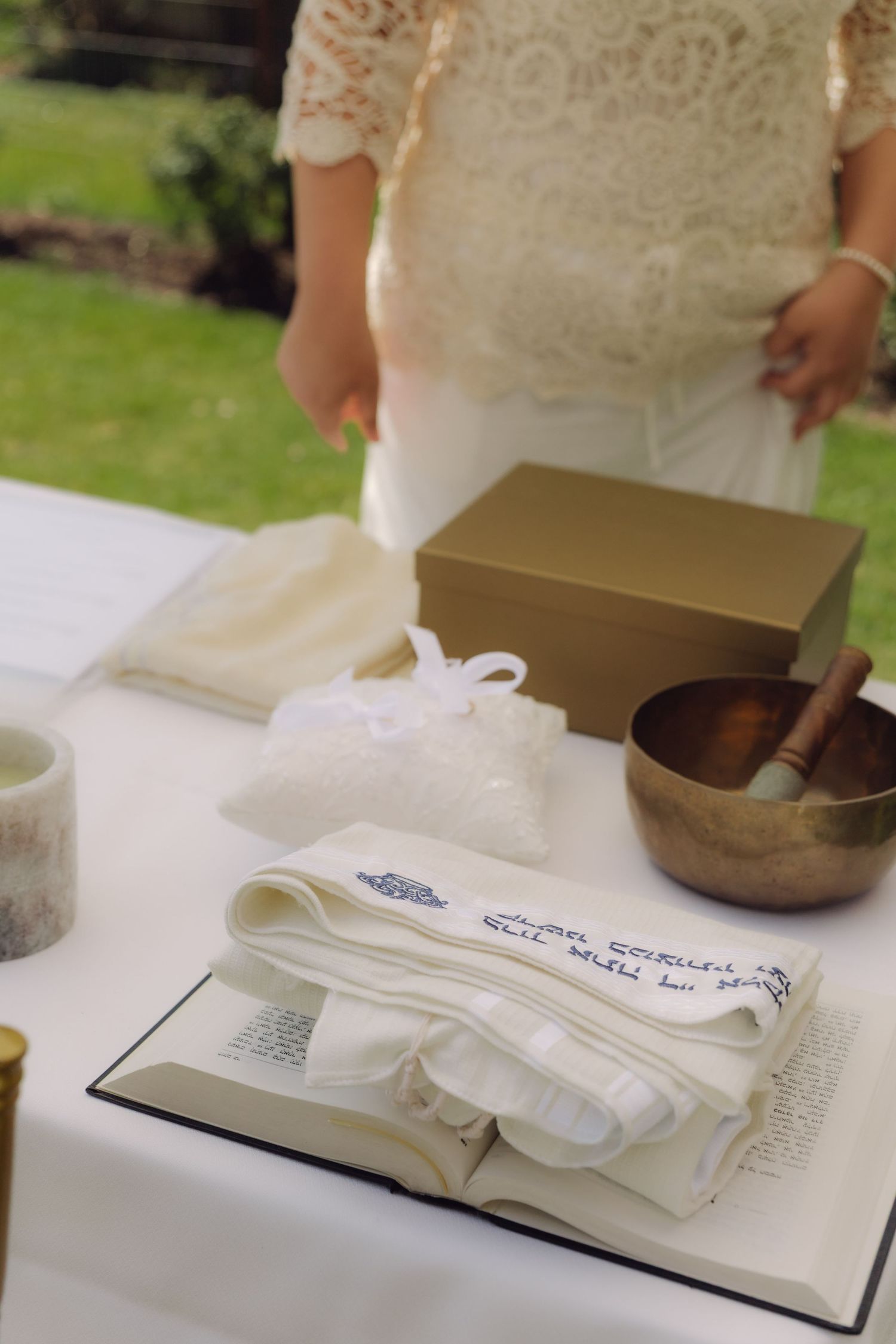 Wedding ceremony altar table with Jewish prayer shawl, open book, singing bowl, and gold box outdoors.
