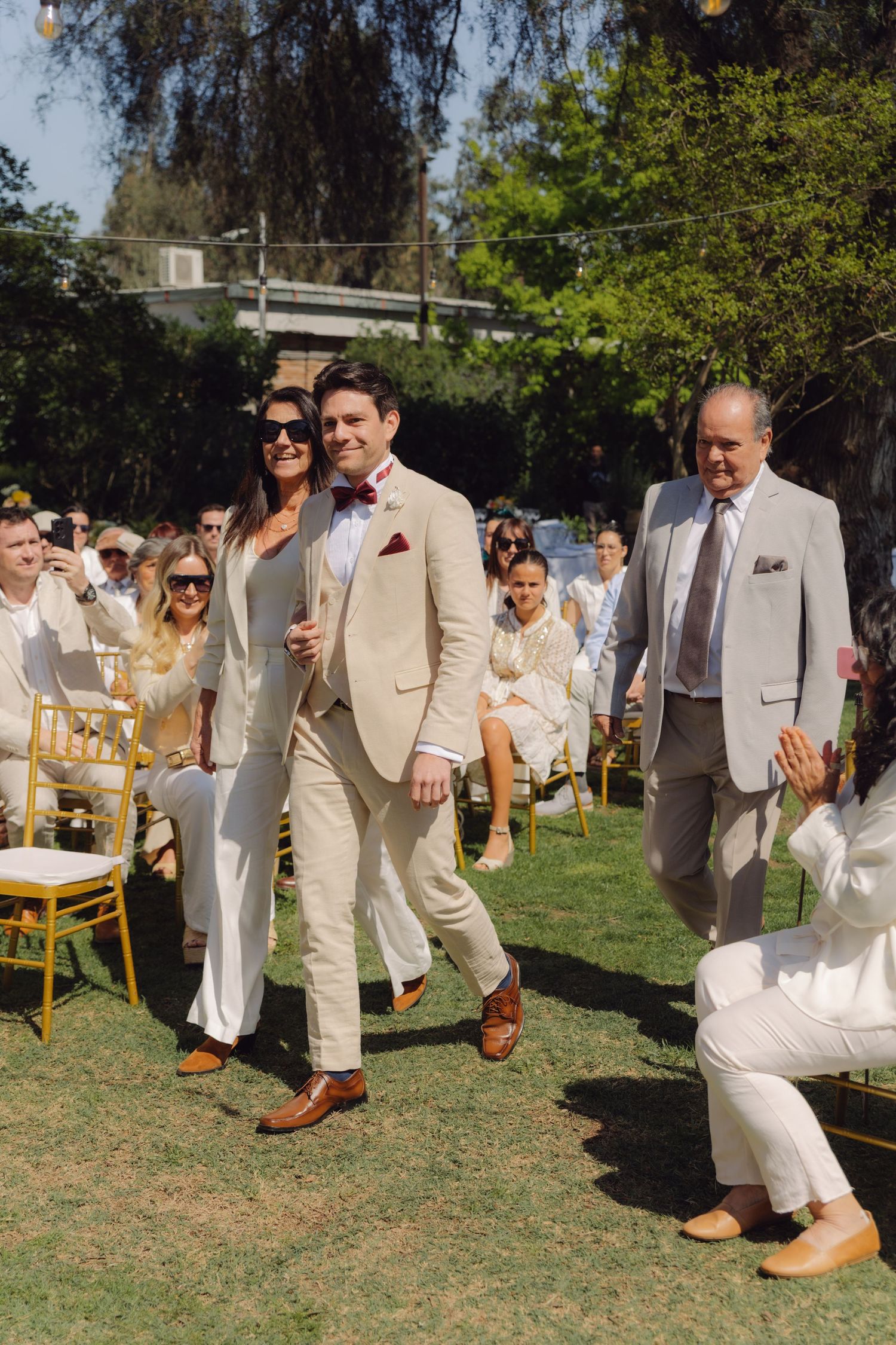 Groom walking down outdoor wedding aisle escorted by parents in beige suits on sunny green lawn.