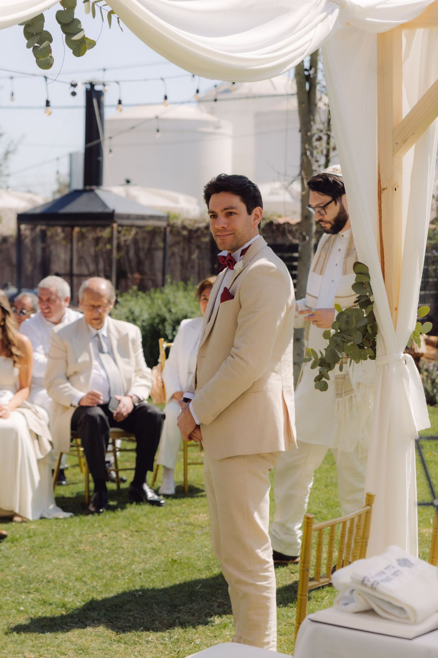 Groom in beige suit with burgundy bow tie standing at outdoor wedding altar with rabbi and guests.