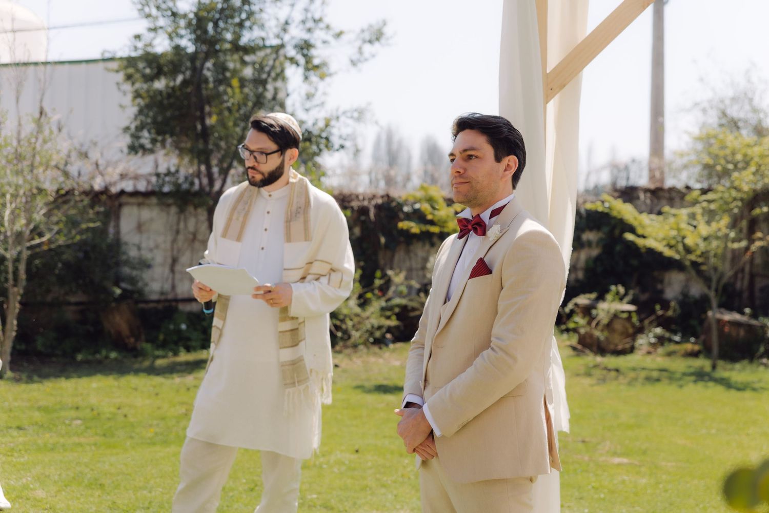 Groom in beige suit with burgundy bow tie standing with rabbi in traditional attire at outdoor wedding.