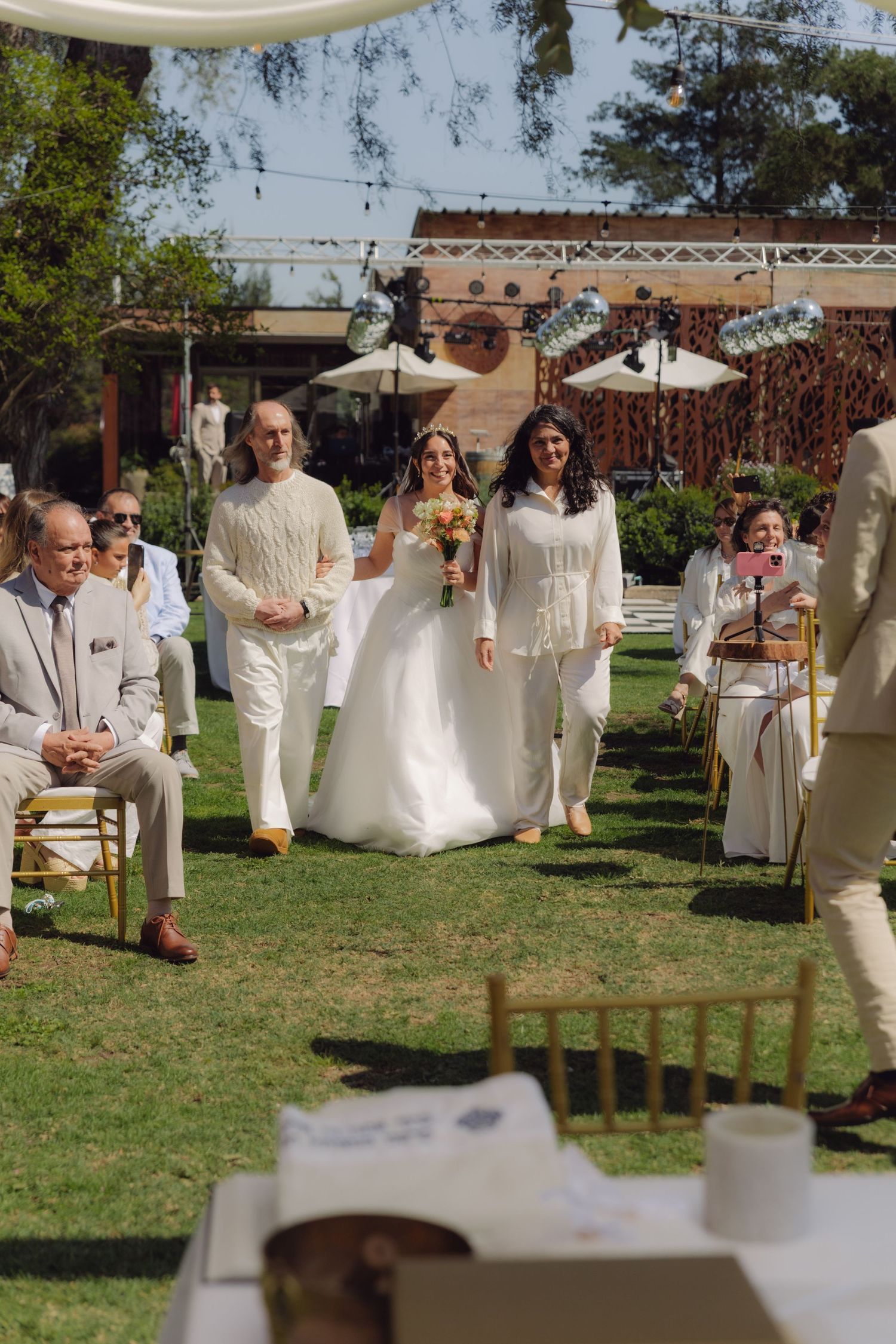 Bride in white ball gown walking down outdoor aisle with family at sunny wedding ceremony.