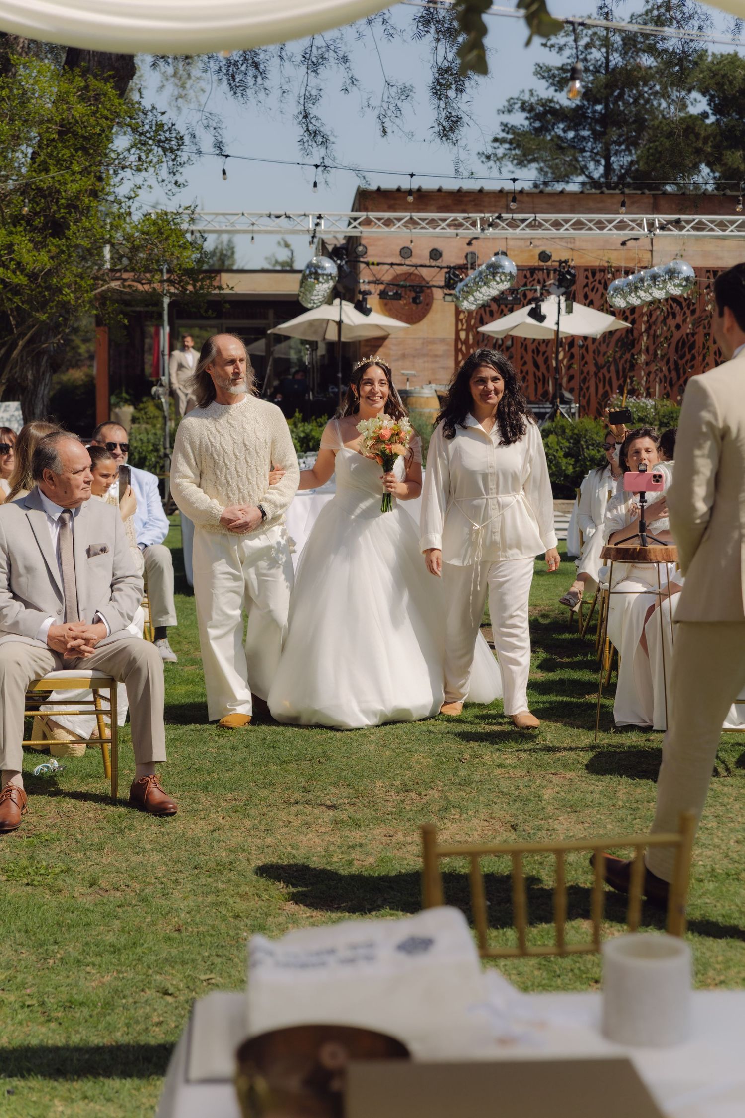 Bride walks down outdoor aisle with family escorts at sunny garden wedding ceremony.