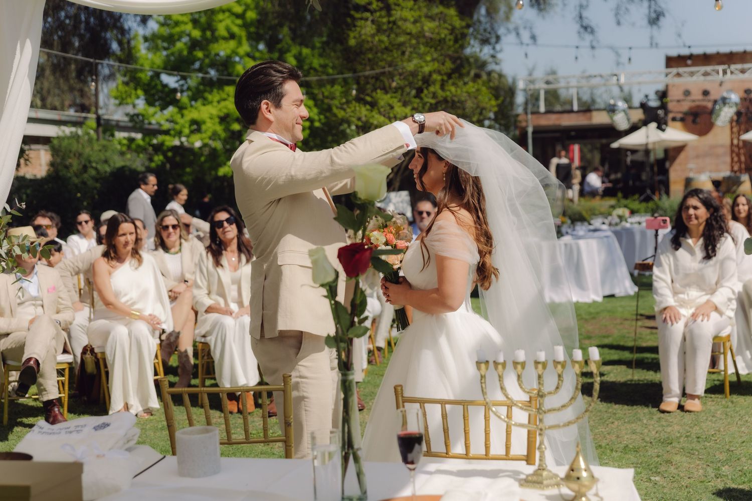 Groom lifts bride's veil during outdoor Jewish wedding ceremony with guests seated in white attire.