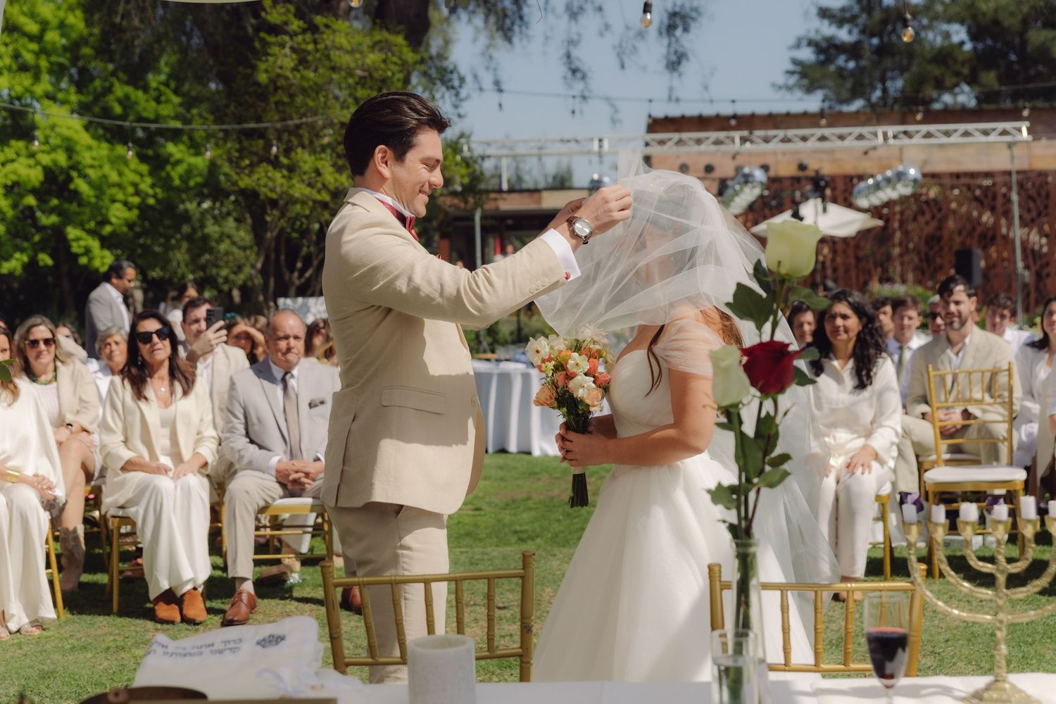 Groom lifts bride's veil during outdoor Jewish wedding ceremony under chuppah.