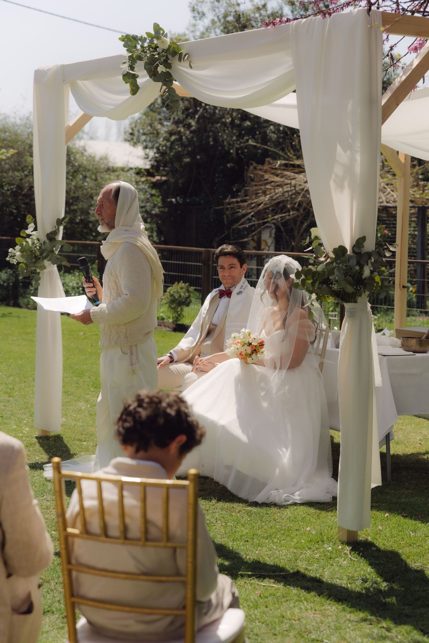 Couple sits under white draped chuppah during Jewish wedding ceremony outdoors.