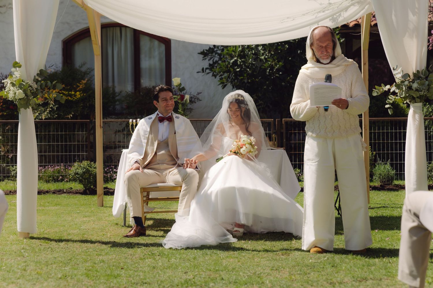 Bride and groom sit under chuppah as officiant reads during elegant outdoor garden wedding ceremony.