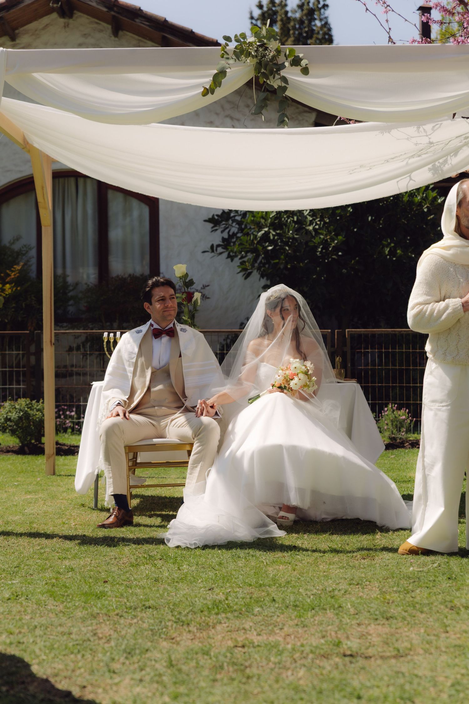 Bride and groom seated under chuppah canopy during outdoor Jewish wedding ceremony.