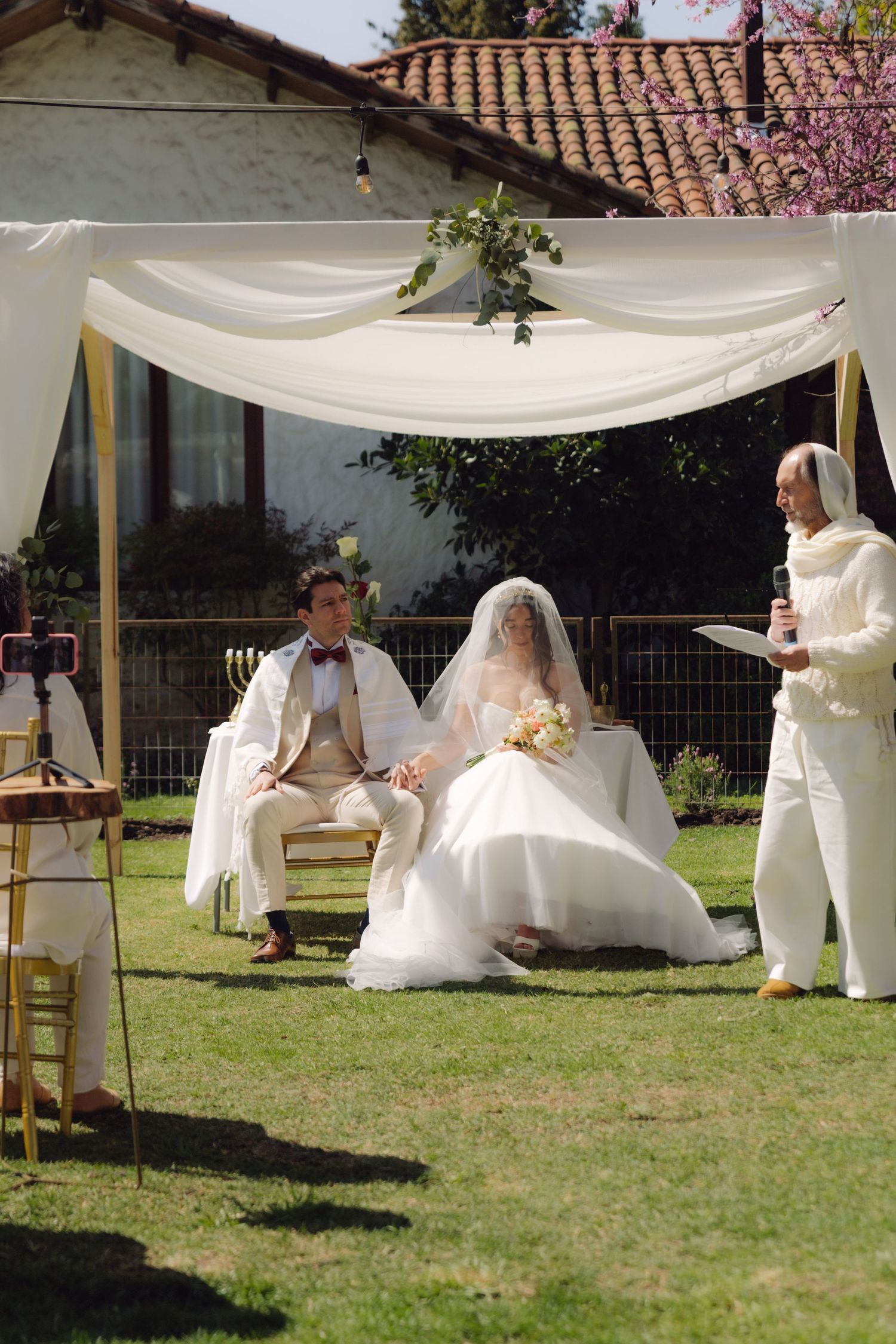 Couple sits under white chuppah as officiant reads during outdoor Jewish wedding.
