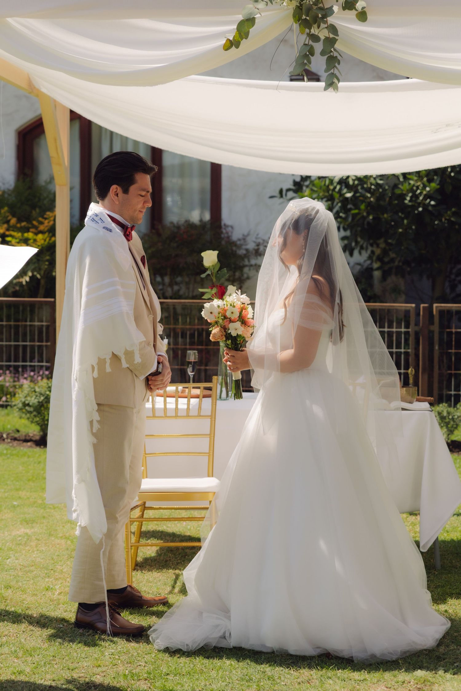 Bride holding bouquet and groom in tallit stand face to face under white chuppah at outdoor wedding.