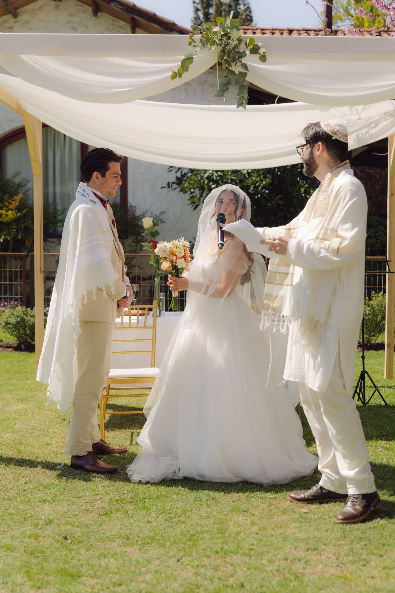 Rabbi officiates Jewish wedding ceremony under chuppah with bride and groom outdoors.