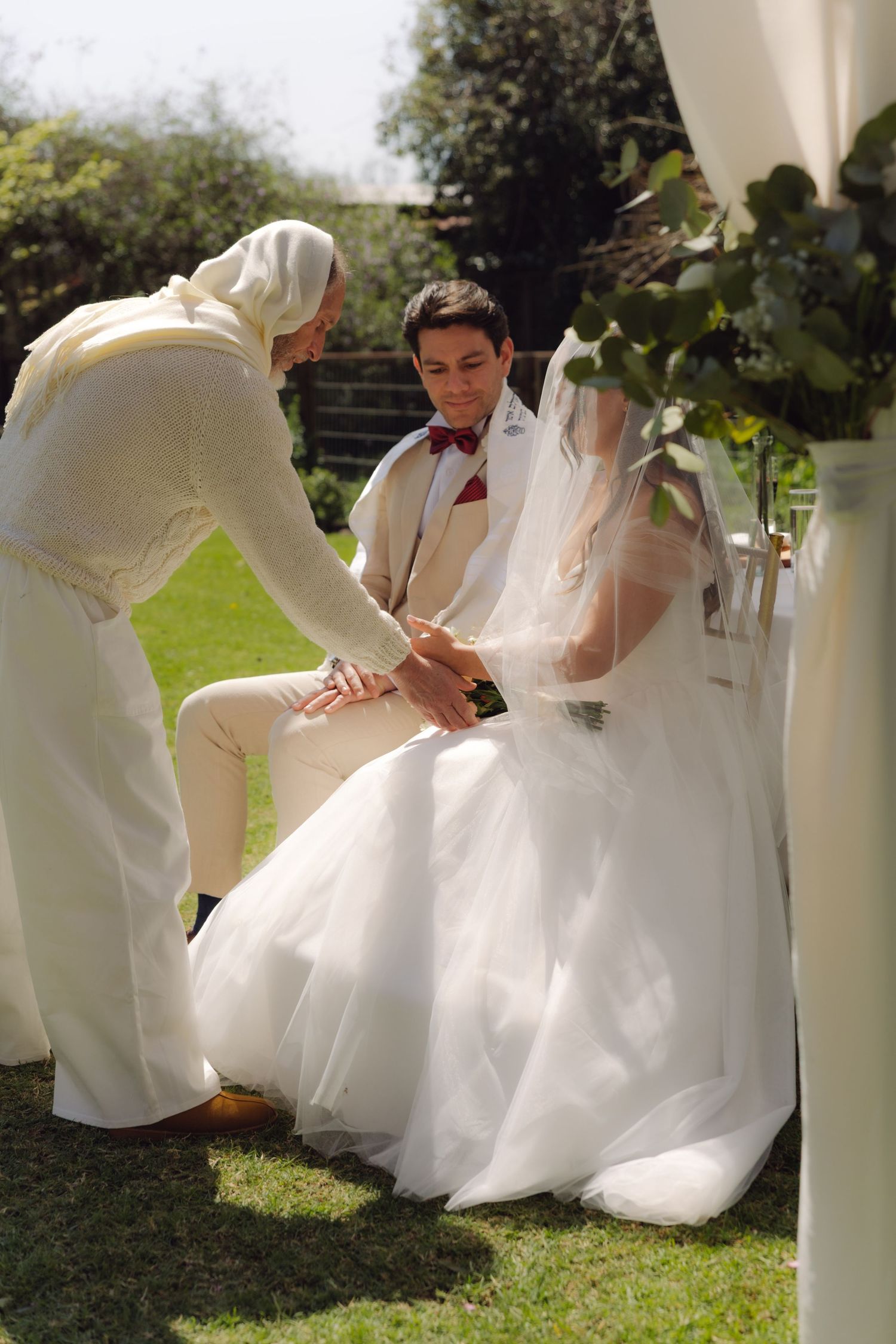 Wedding officiant performs hand blessing ritual with couple under outdoor chuppah.