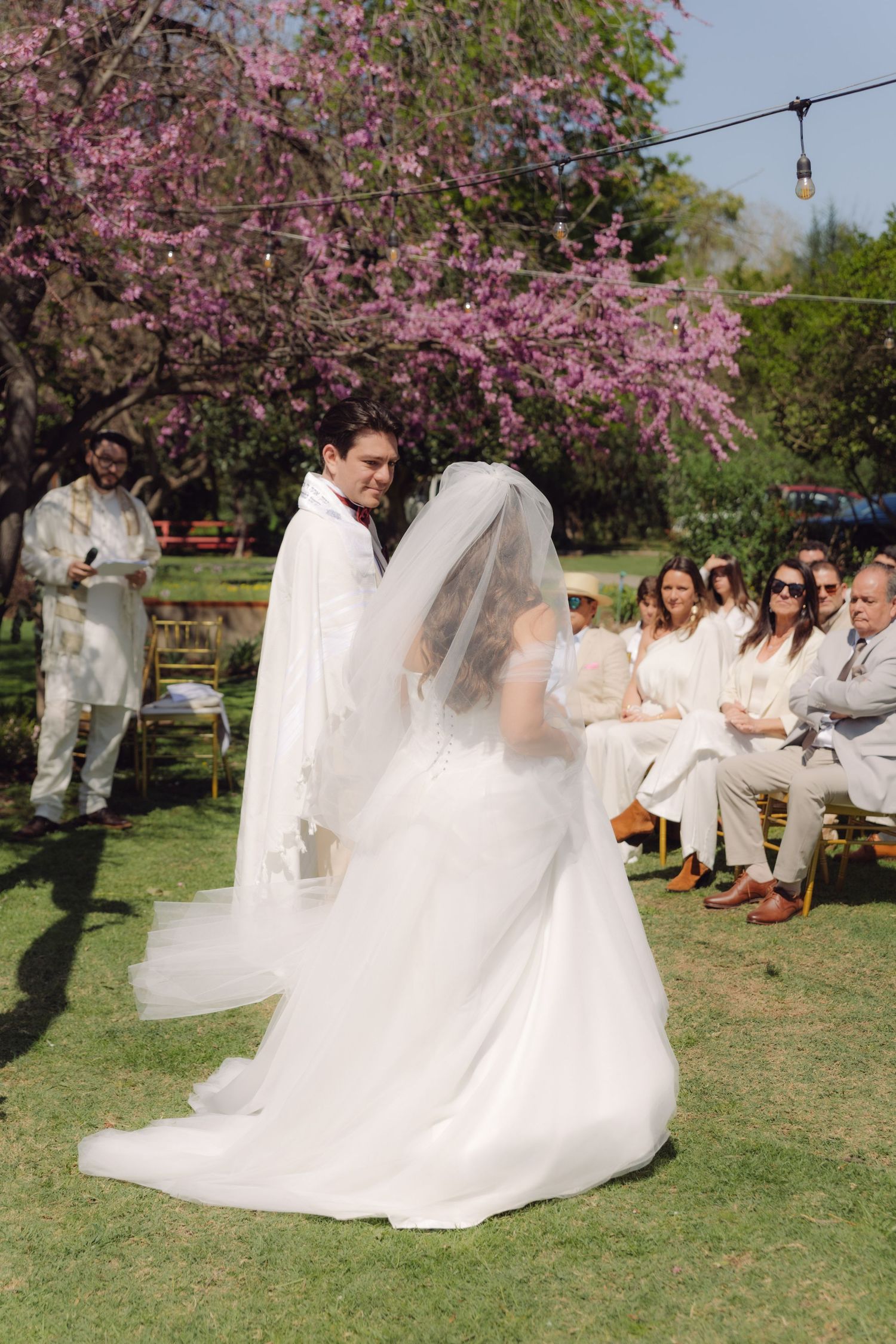 Groom lifts bride's veil during an outdoor Jewish wedding ceremony beneath a blooming pink tree.