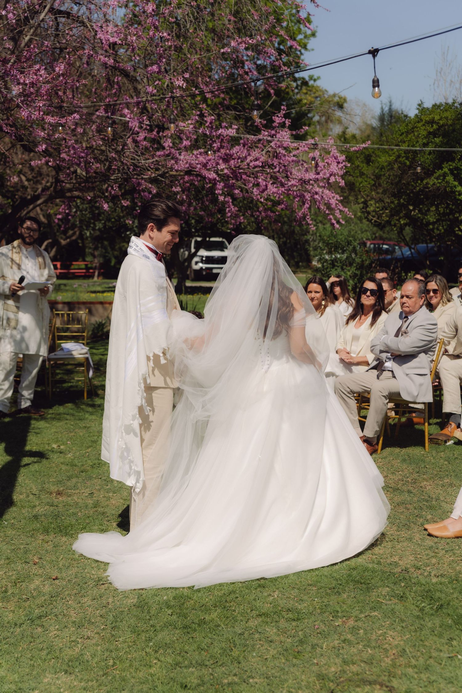Bride in white gown and groom in tallit stand before seated guests under pink blossoming tree.