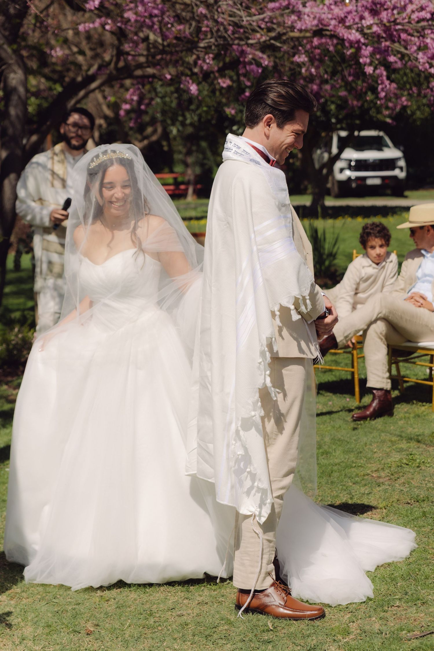Smiling bride in white gown walks with groom wearing tallit under pink flowering tree at wedding.
