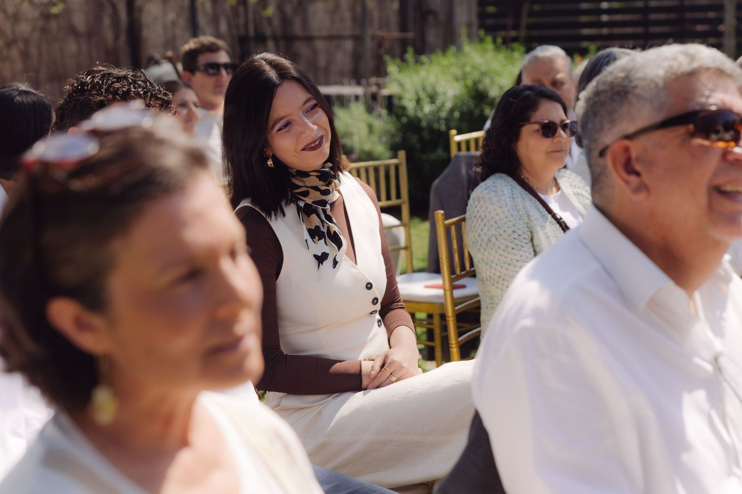 Smiling wedding guests seated in gold chairs enjoy an outdoor ceremony on a sunny day.