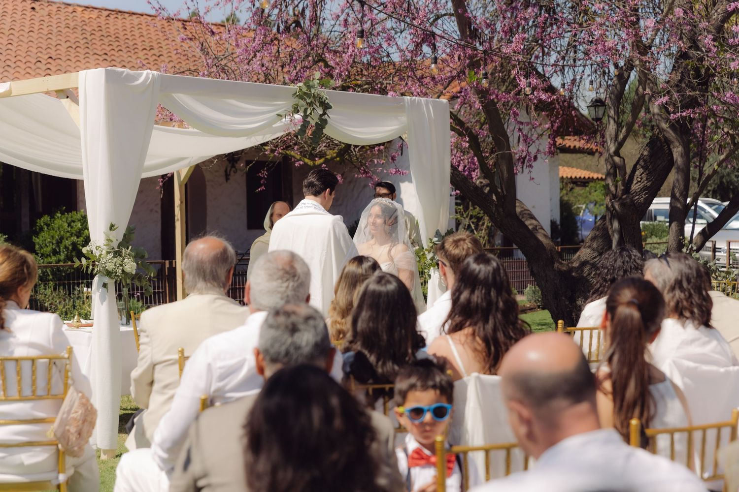 Guests watch bride and groom exchange vows under a white floral chuppah at an outdoor wedding.
