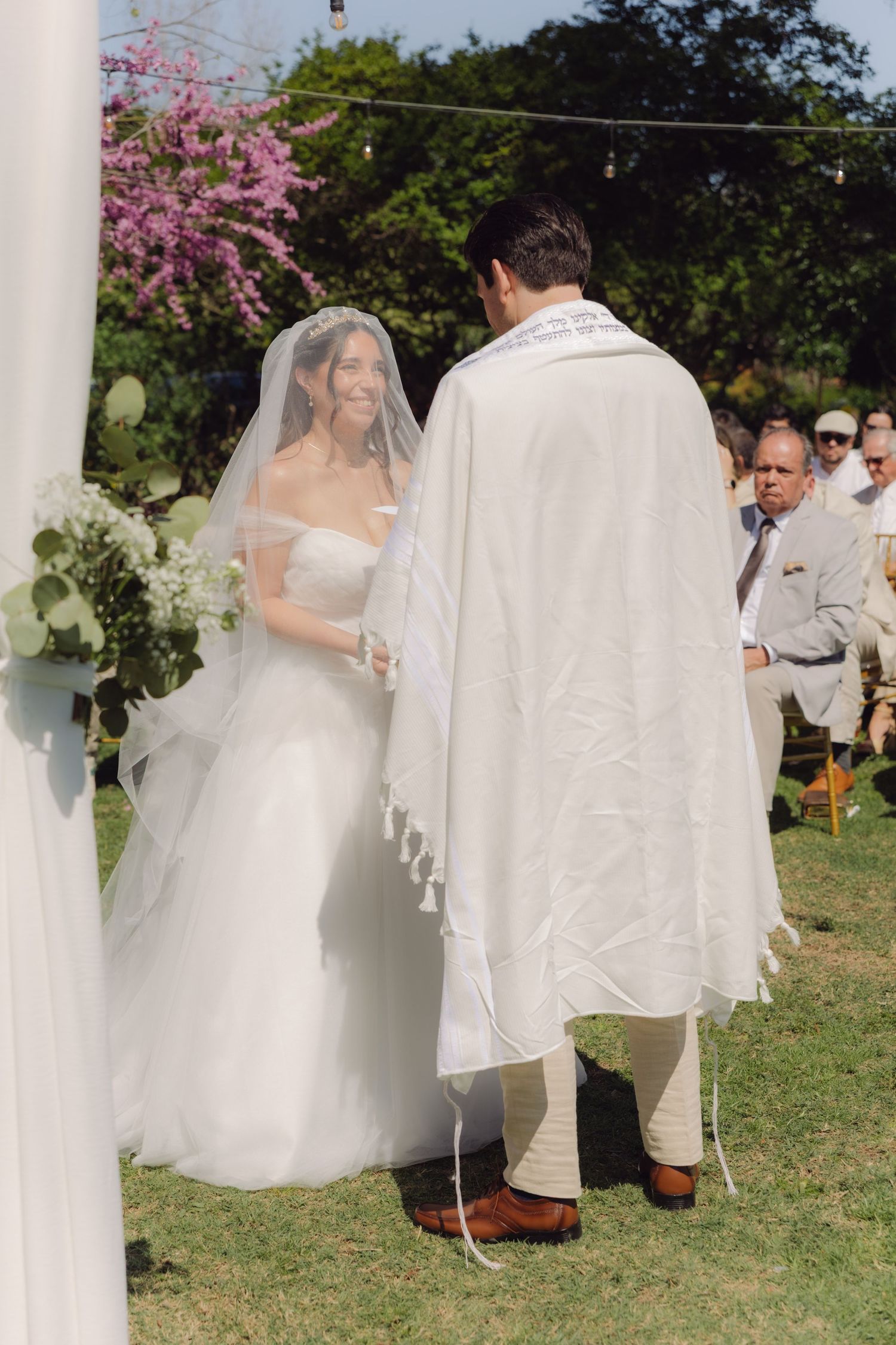 Smiling bride in white gown holds flowers facing groom wearing a tallit at outdoor ceremony.