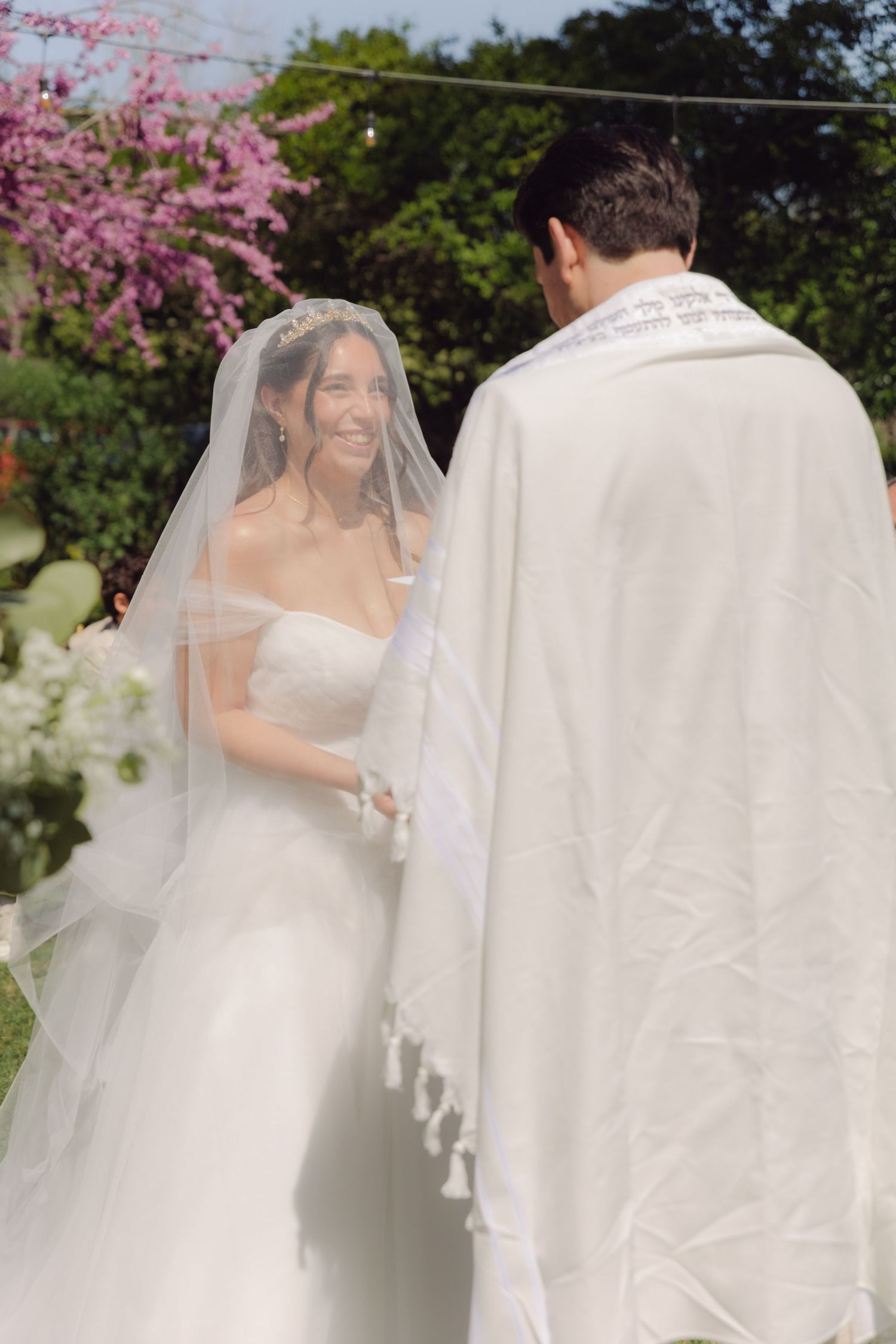 Radiant bride smiles at groom wearing a white tallit during an outdoor garden wedding ceremony.