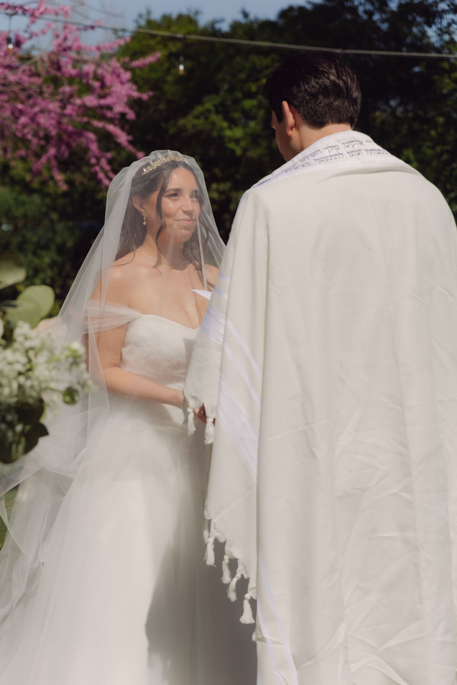 Radiant bride in white gown and veil exchanges vows with groom in tallit at outdoor ceremony.