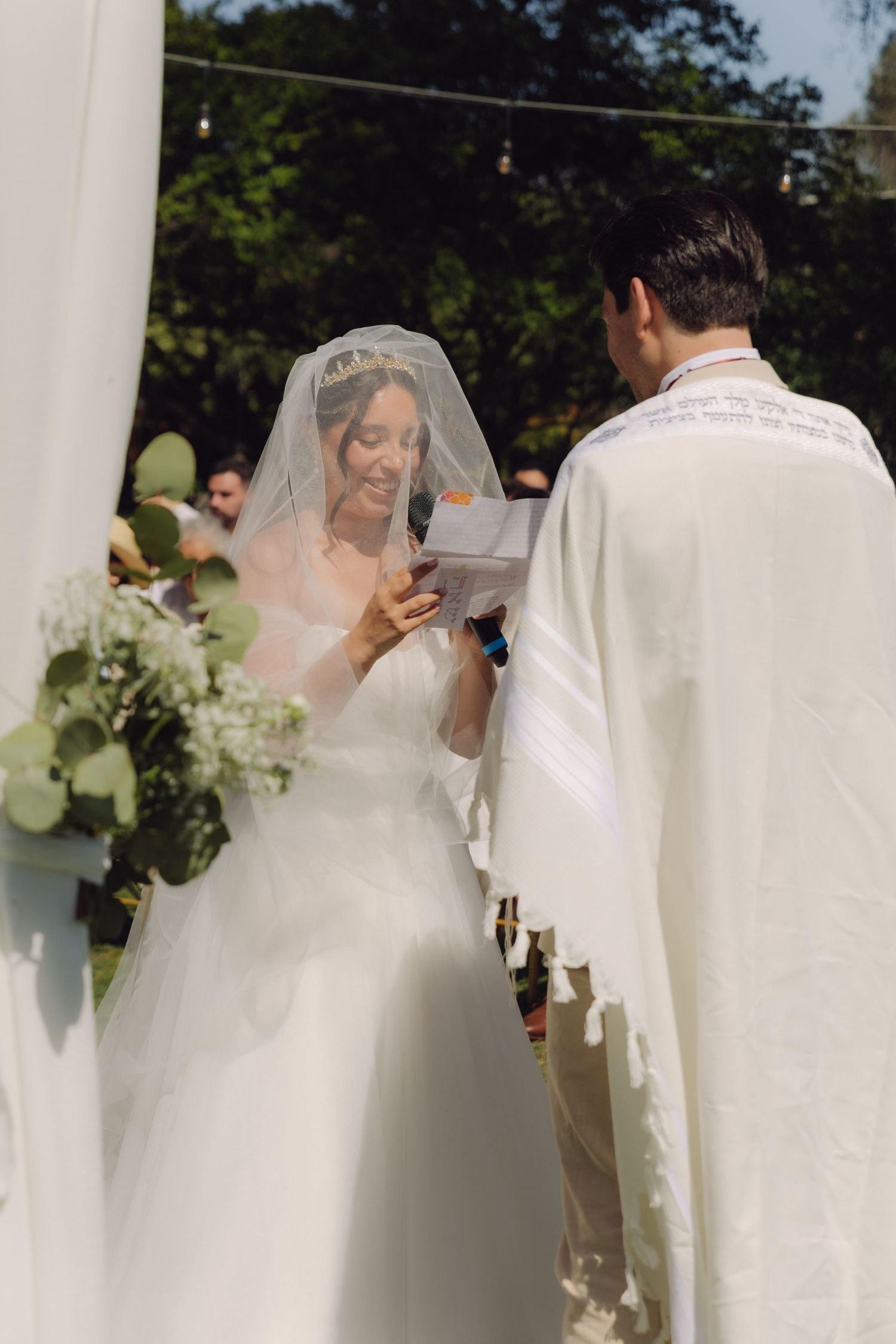 Bride reads vows from card while groom in white tallit listens at sunlit outdoor wedding ceremony.