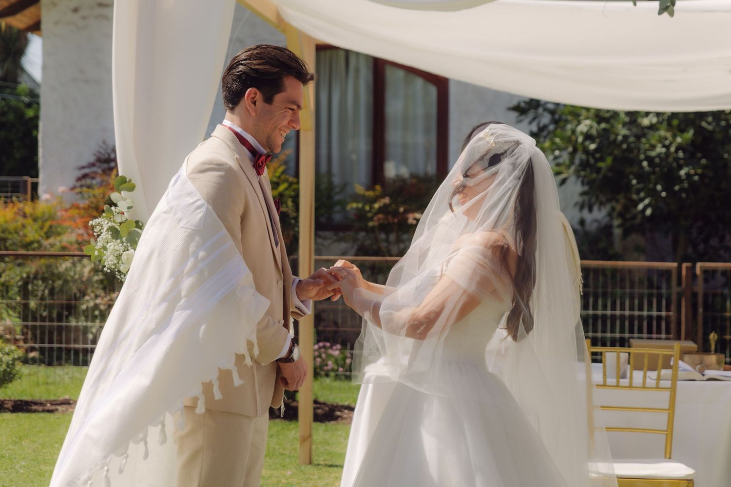 Groom placing wedding ring on brides finger at outdoor altar, bride wearing veil under a white chuppah canopy.