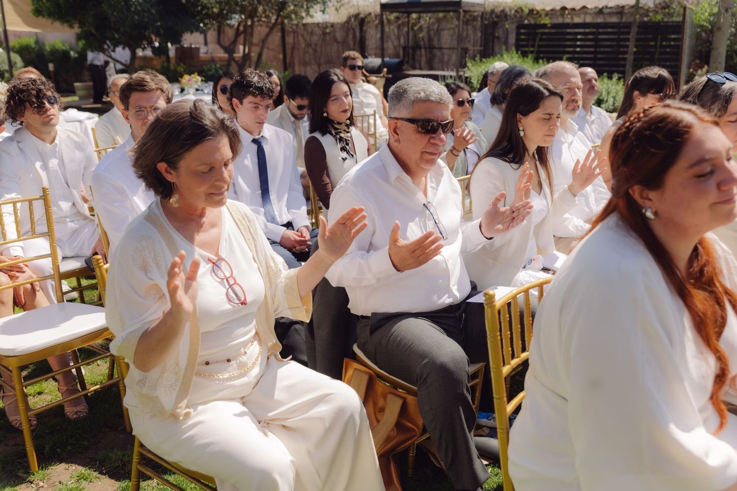 Wedding guests dressed in white applaud during outdoor garden ceremony on sunny day.