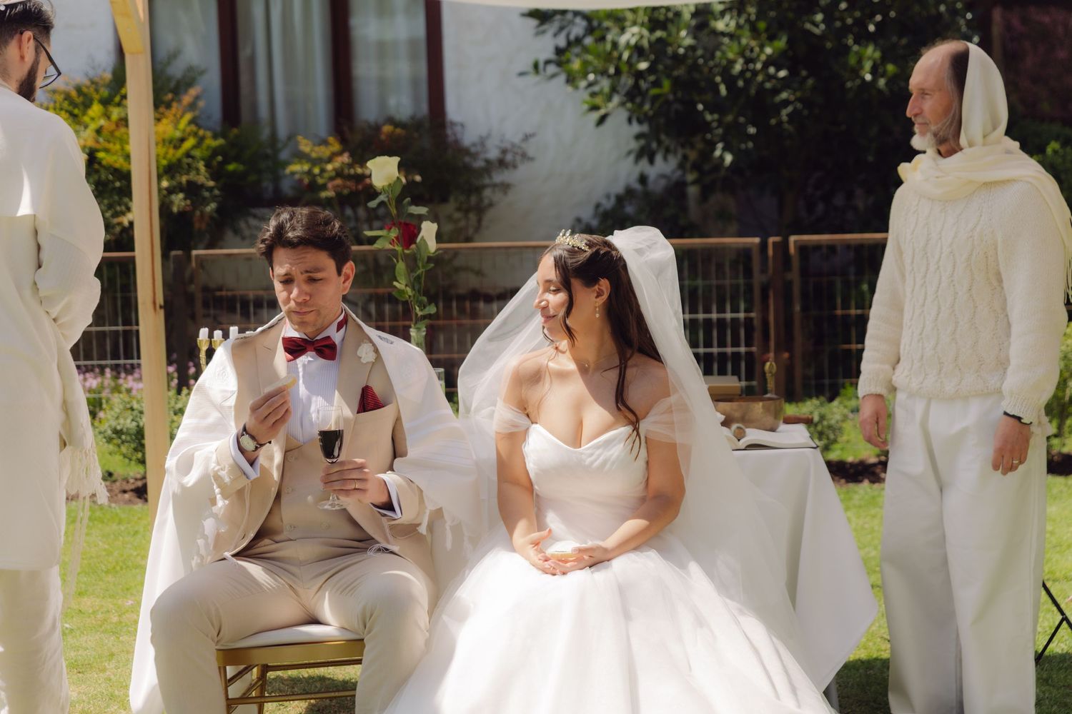 Bride and groom seated during outdoor wedding ceremony, groom reading vows in beige suit with burgundy bow tie.