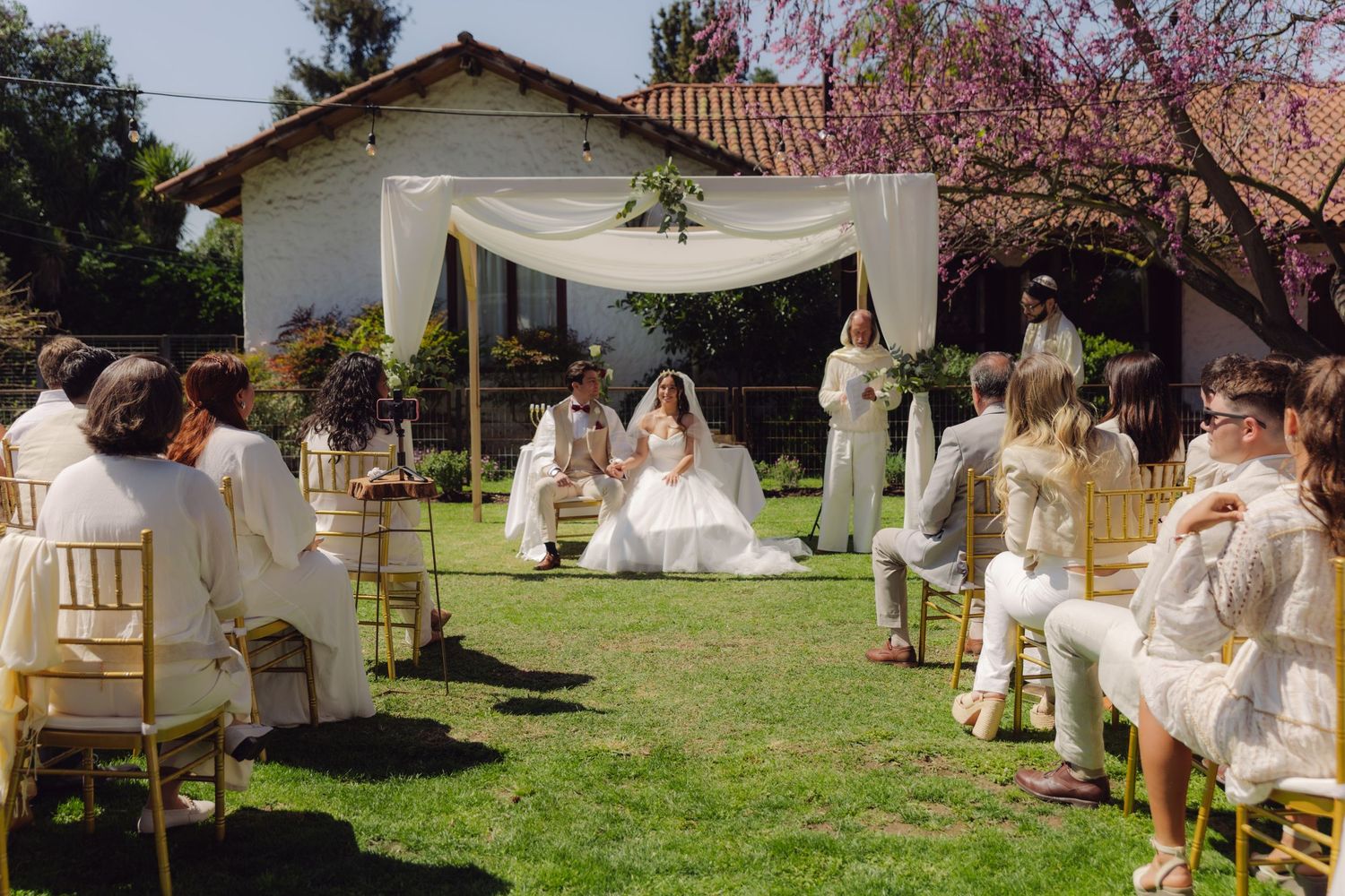 Elegant outdoor wedding ceremony with white floral arch, bride and groom seated surrounded by guests in white attire.