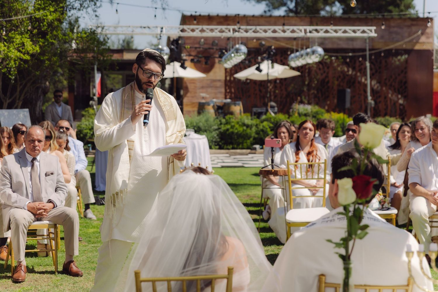 Wedding officiant speaking into microphone during outdoor garden ceremony with seated guests in white and beige outfits.