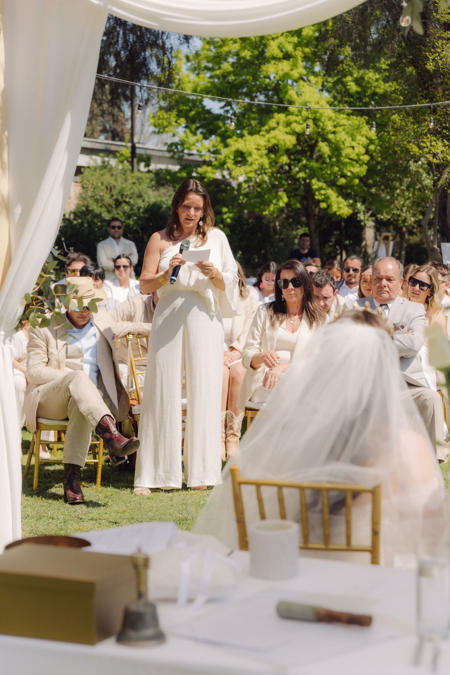 Woman in white jumpsuit delivering a reading during an outdoor wedding ceremony with guests seated on gold chairs.