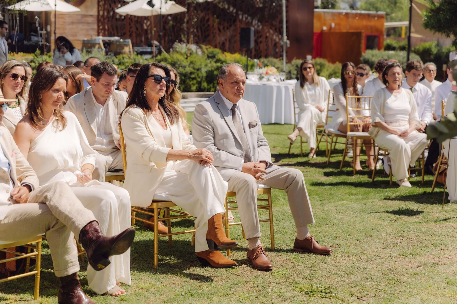 Wedding guests dressed in white and beige seated on gold chairs during an elegant outdoor garden ceremony.