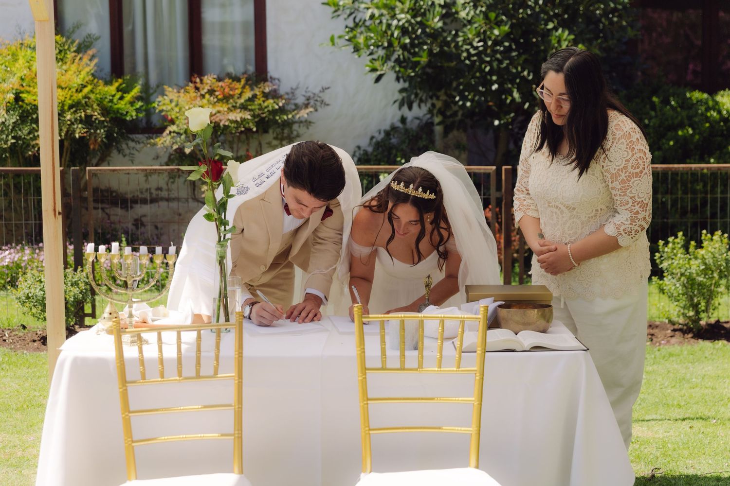 Bride and groom signing wedding documents at a white table with gold chairs, officiant standing beside them.