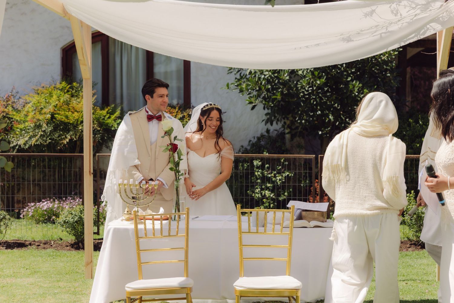 Bride in strapless white gown and groom in beige suit standing together under a draped wedding canopy outdoors.