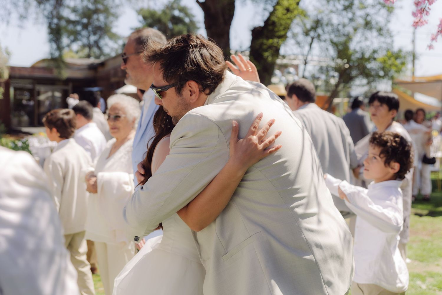 Newlywed couple sharing a warm embrace with guests celebrating after an outdoor garden wedding ceremony.