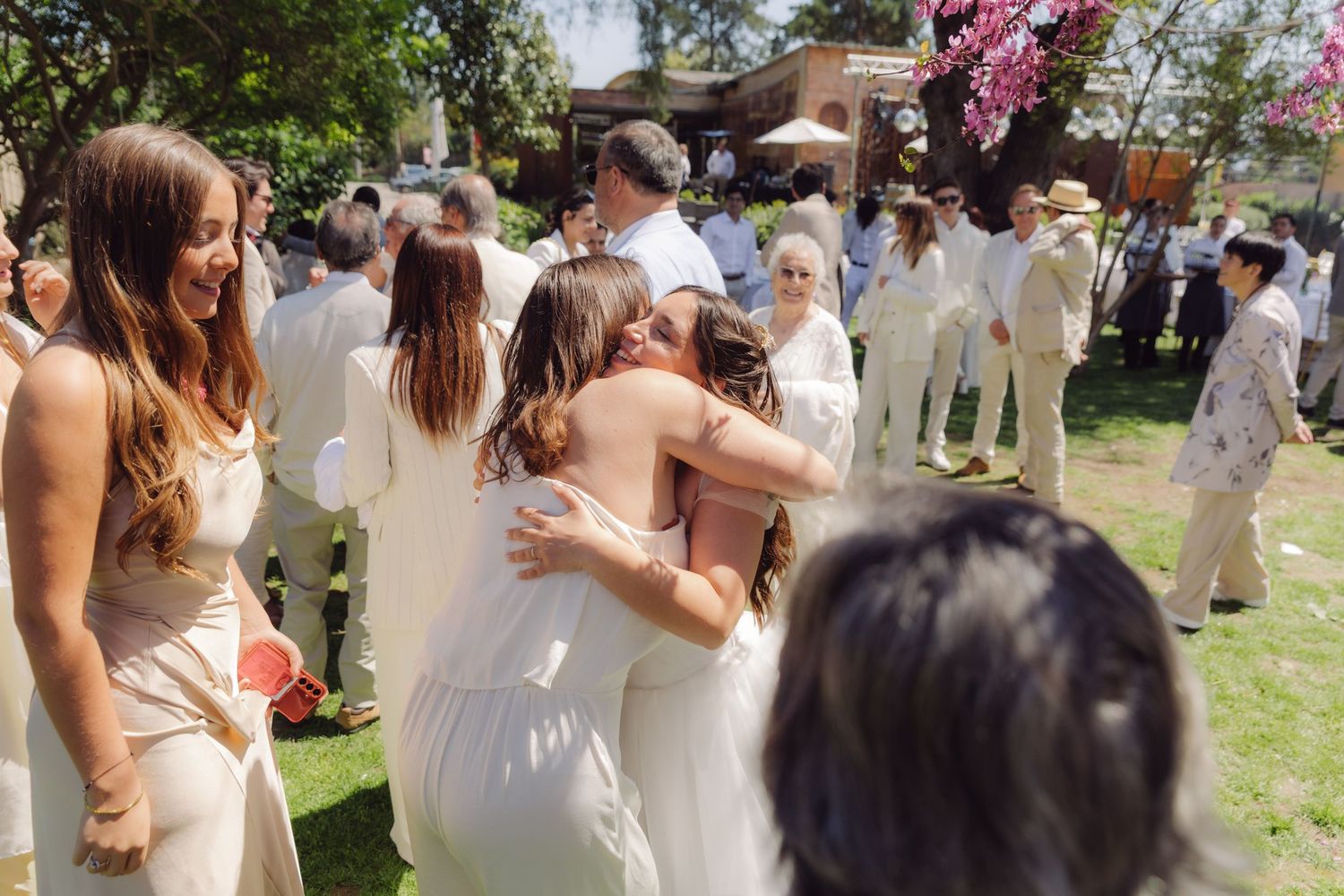 Bride in white gown hugging a guest while smiling friends celebrate after an outdoor wedding ceremony.