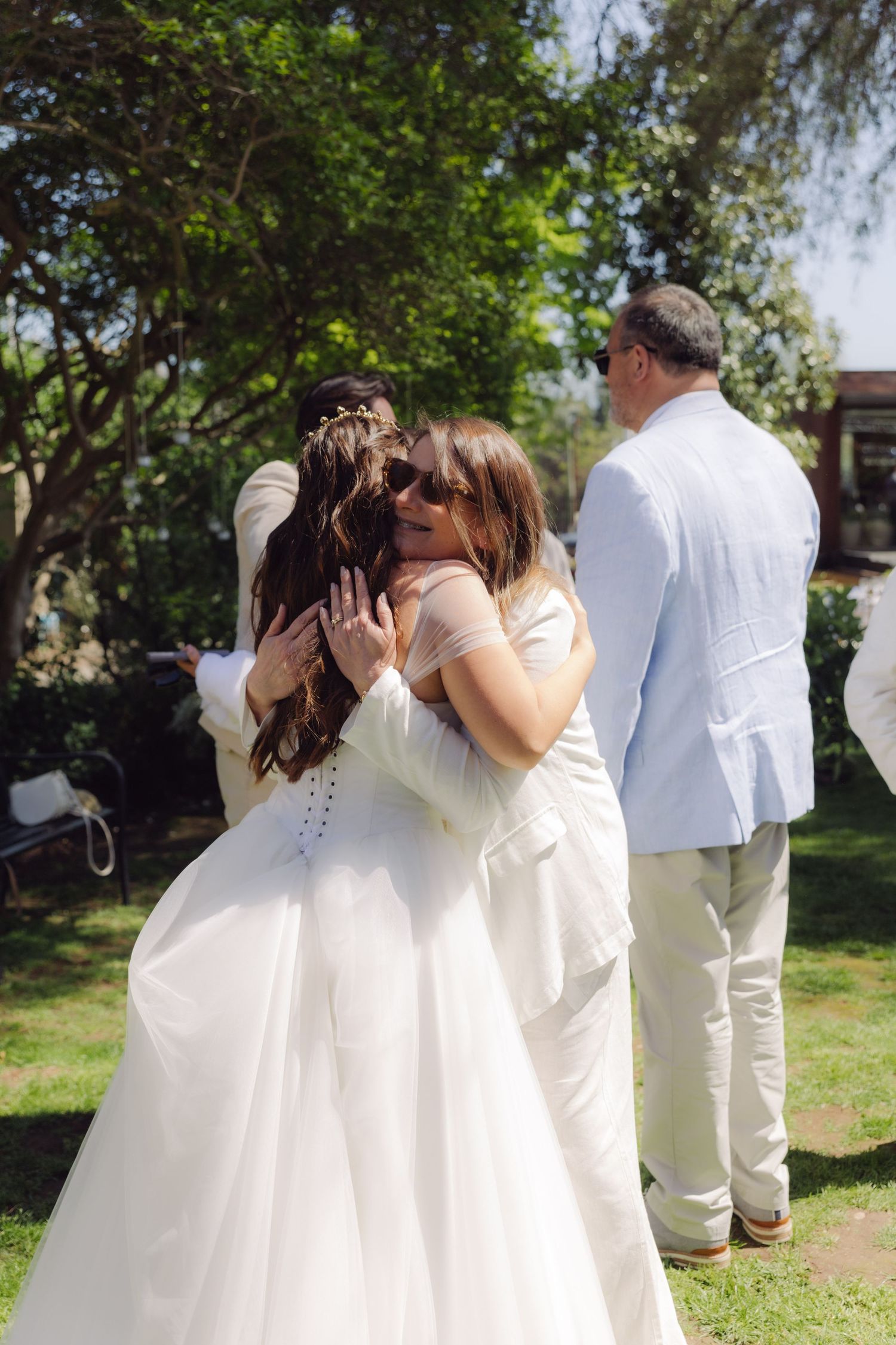 Bride in white gown receiving a warm hug from a guest in sunglasses during outdoor post-ceremony celebrations.