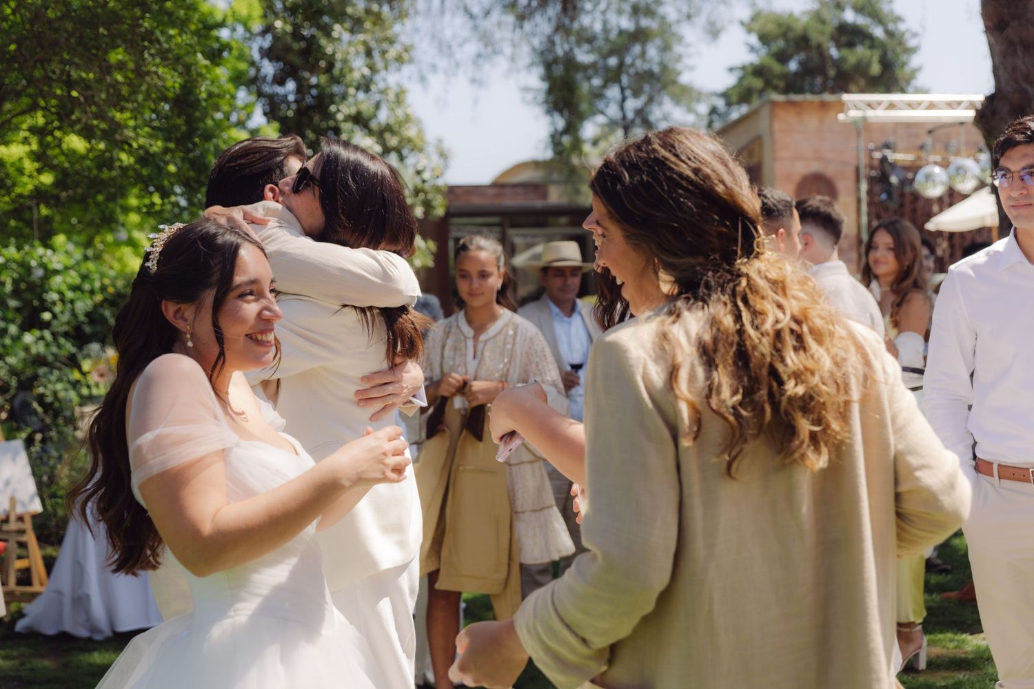 Bride hugging a guest while smiling friends gather to celebrate after a sunny outdoor garden wedding ceremony.