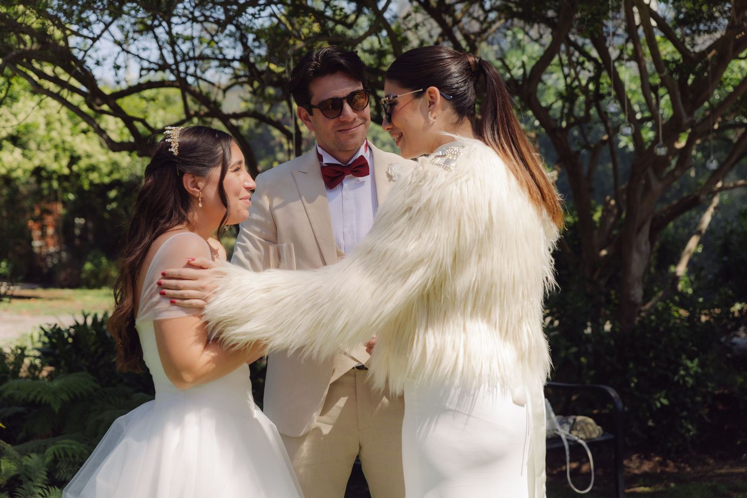 Bride and groom sharing a joyful moment with a guest in a white fur jacket at an outdoor wedding.