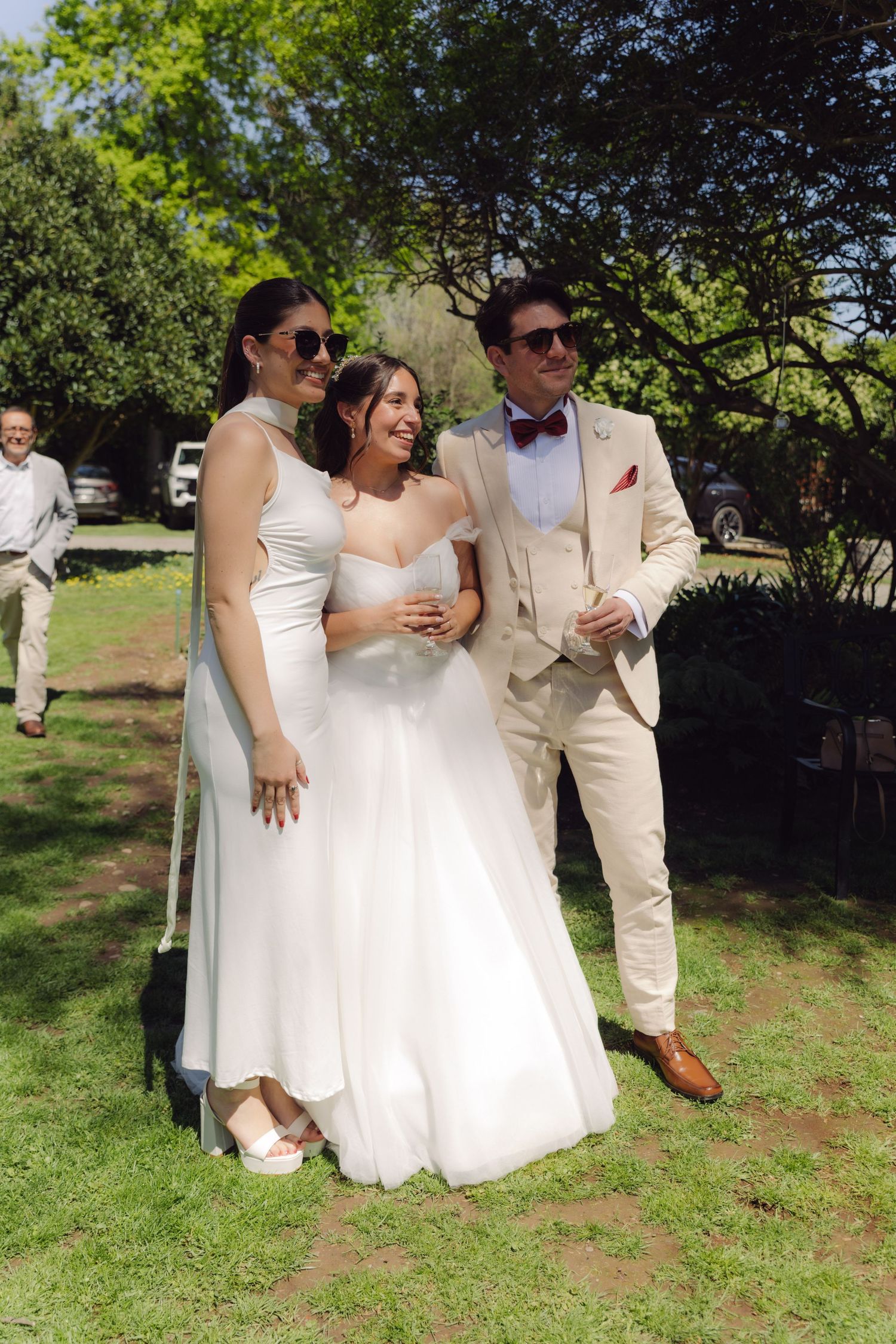 Bride in white gown and groom in beige suit posing with a female guest at an outdoor wedding.